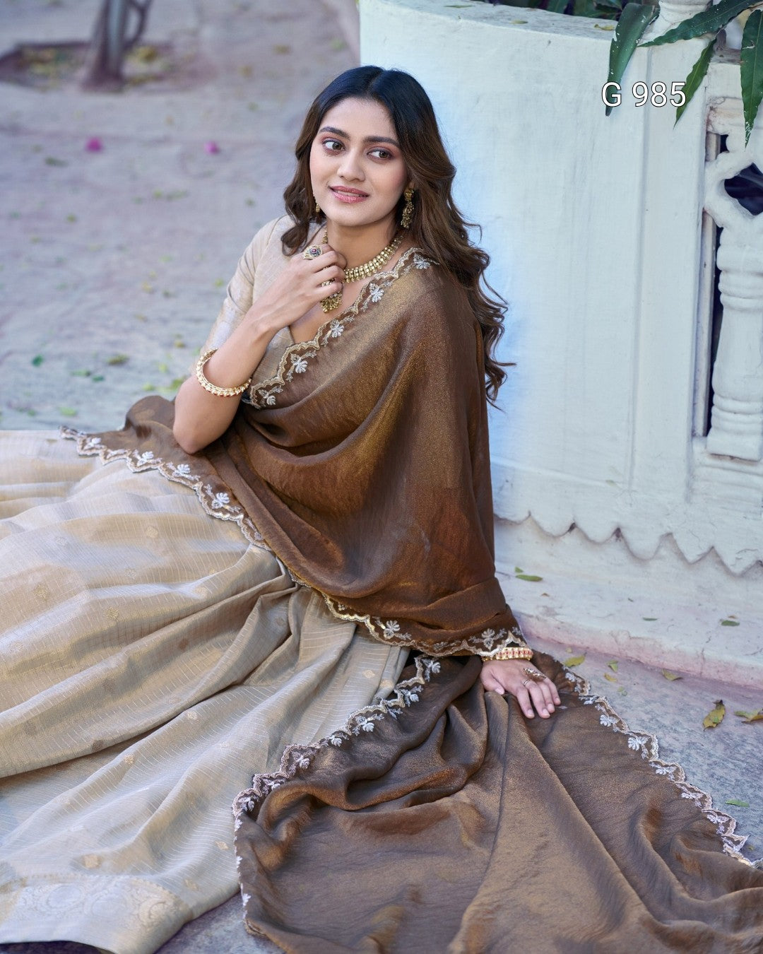 A woman wearing a traditional Indian outfit with a brown shawl draped over a beige kurta and matching skirt, seated outdoors against a white wall.