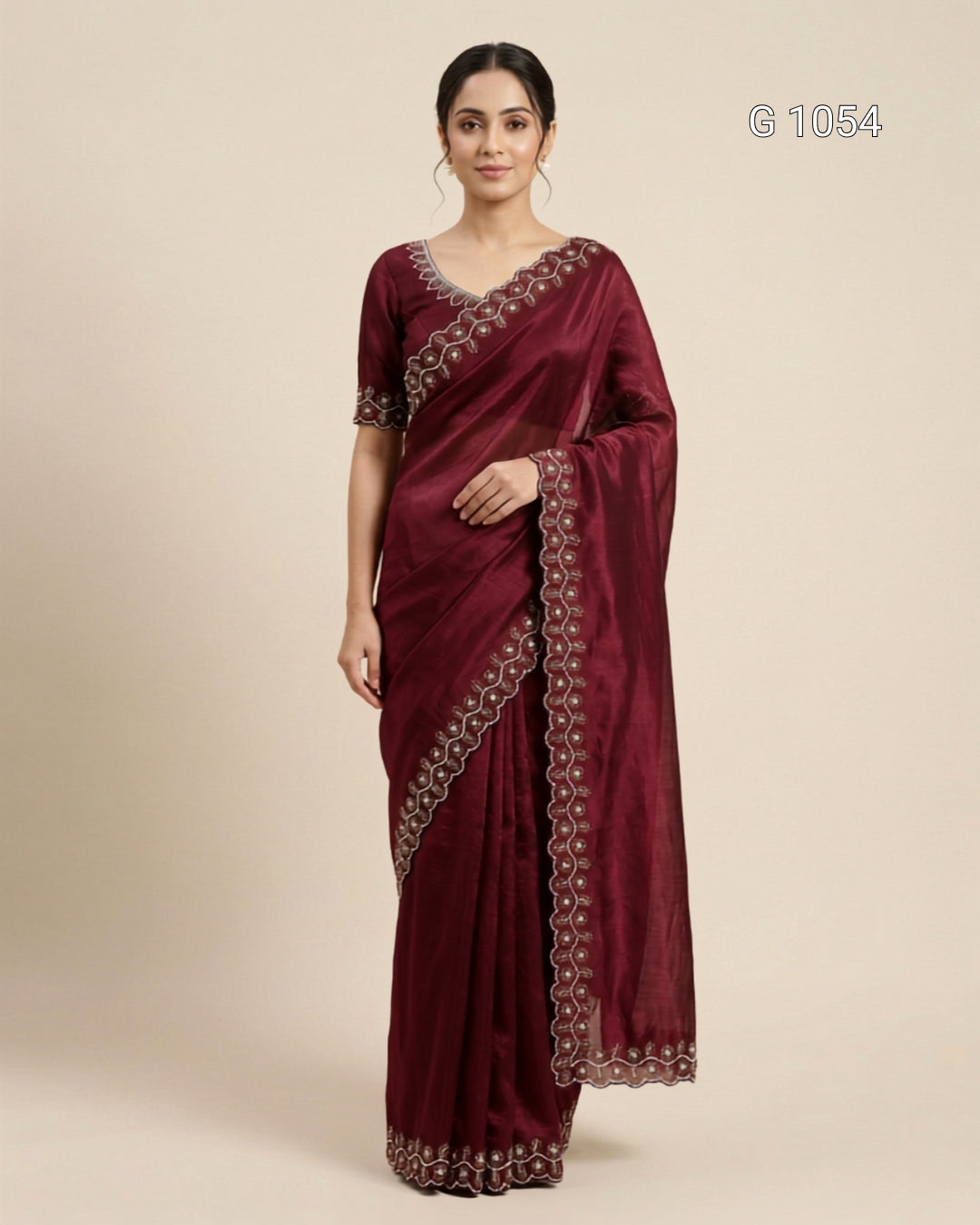 A woman modeling a deep maroon silk saree with intricate silver and gold embroidered borders, paired with a matching embroidered blouse, set against a plain beige background.
