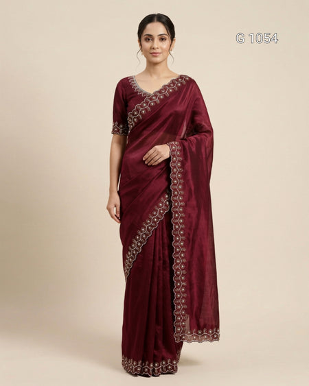 A woman modeling a deep maroon silk saree with intricate silver and gold embroidered borders, paired with a matching embroidered blouse, set against a plain beige background.