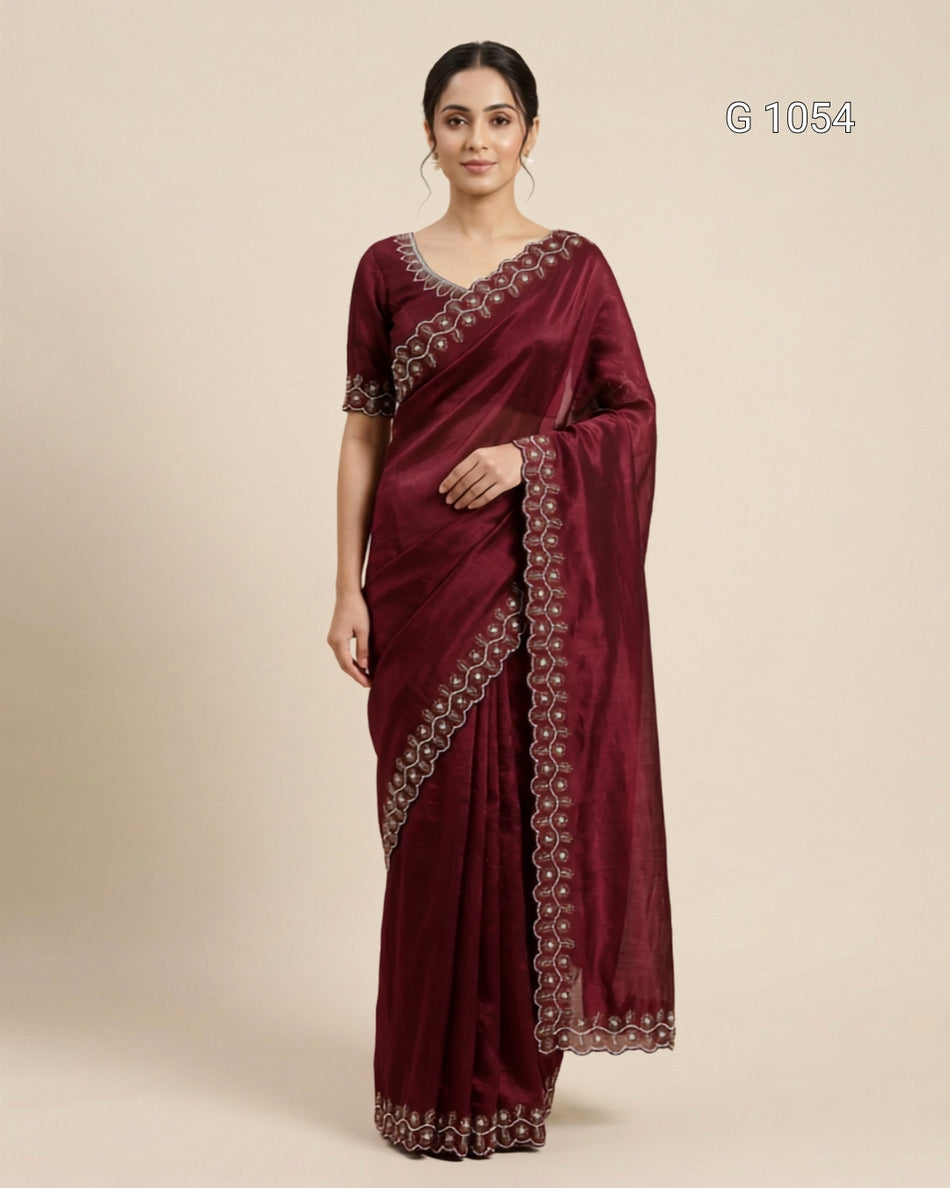 A woman modeling a deep maroon silk saree with intricate silver and gold embroidered borders, paired with a matching embroidered blouse, set against a plain beige background.