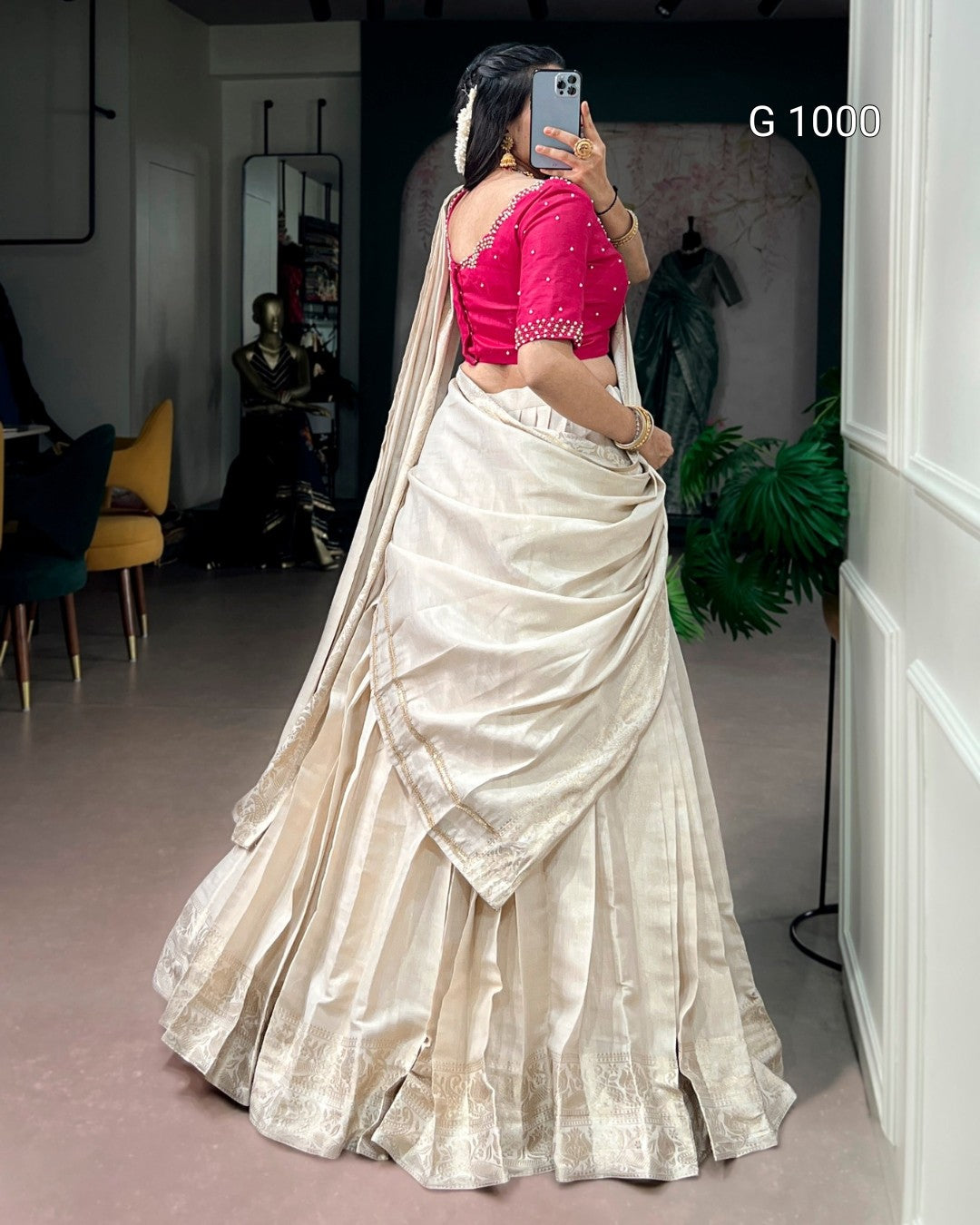 A woman takes a mirror selfie in a boutique wearing a traditional Indian saree. The saree consists of a voluminous cream-colored skirt with a subtle sheen and border, paired with a bright pink embroidered blouse.