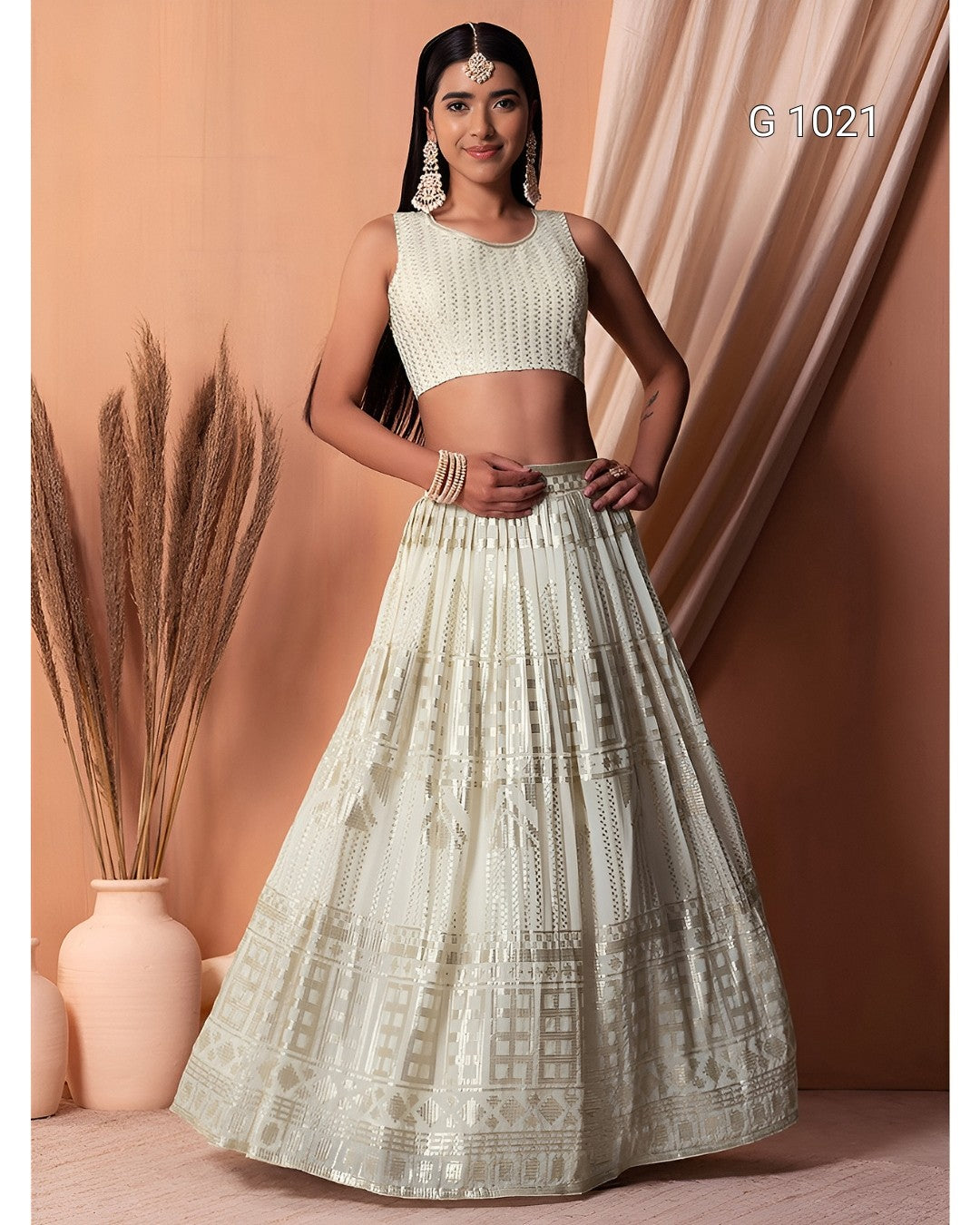 A woman models a traditional Indian outfit consisting of a sleeveless white embroidered crop top and a matching flared skirt with geometric patterns, set against a warm peach-colored backdrop with decorative dried grass in a vase.