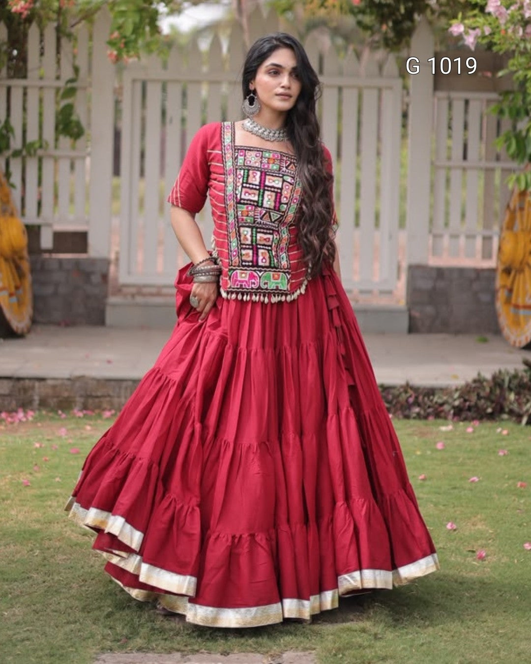 A woman modeling a traditional Indian red lehenga choli set with intricate multicolor embroidery on the chest and a tiered skirt with gold borders, set against a garden background.