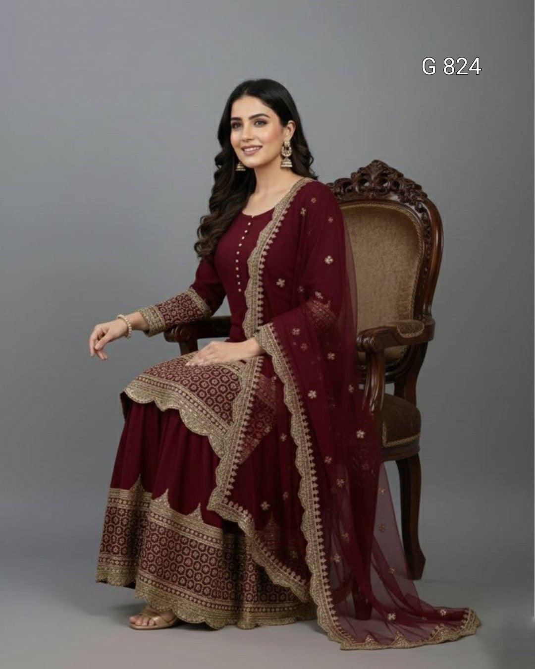 Woman in a maroon traditional outfit with gold embroidery sitting on a wooden chair against a gray background.