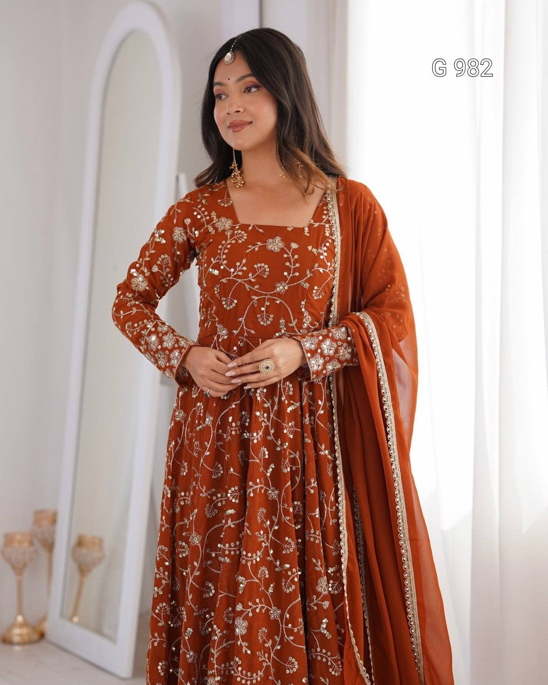 A woman modeling a rust-orange Anarkali gown with white floral embroidery, paired with a matching dupatta, in a bright indoor setting.