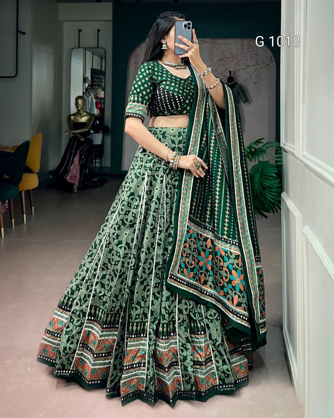 A woman takes a mirror selfie wearing a green printed traditional lehenga choli set with a matching dupatta. The outfit features intricate black and orange geometric patterns along the borders of the skirt and dupatta.