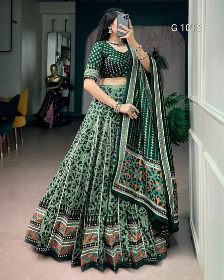 A woman takes a mirror selfie wearing a green printed traditional lehenga choli set with a matching dupatta. The outfit features intricate black and orange geometric patterns along the borders of the skirt and dupatta.
