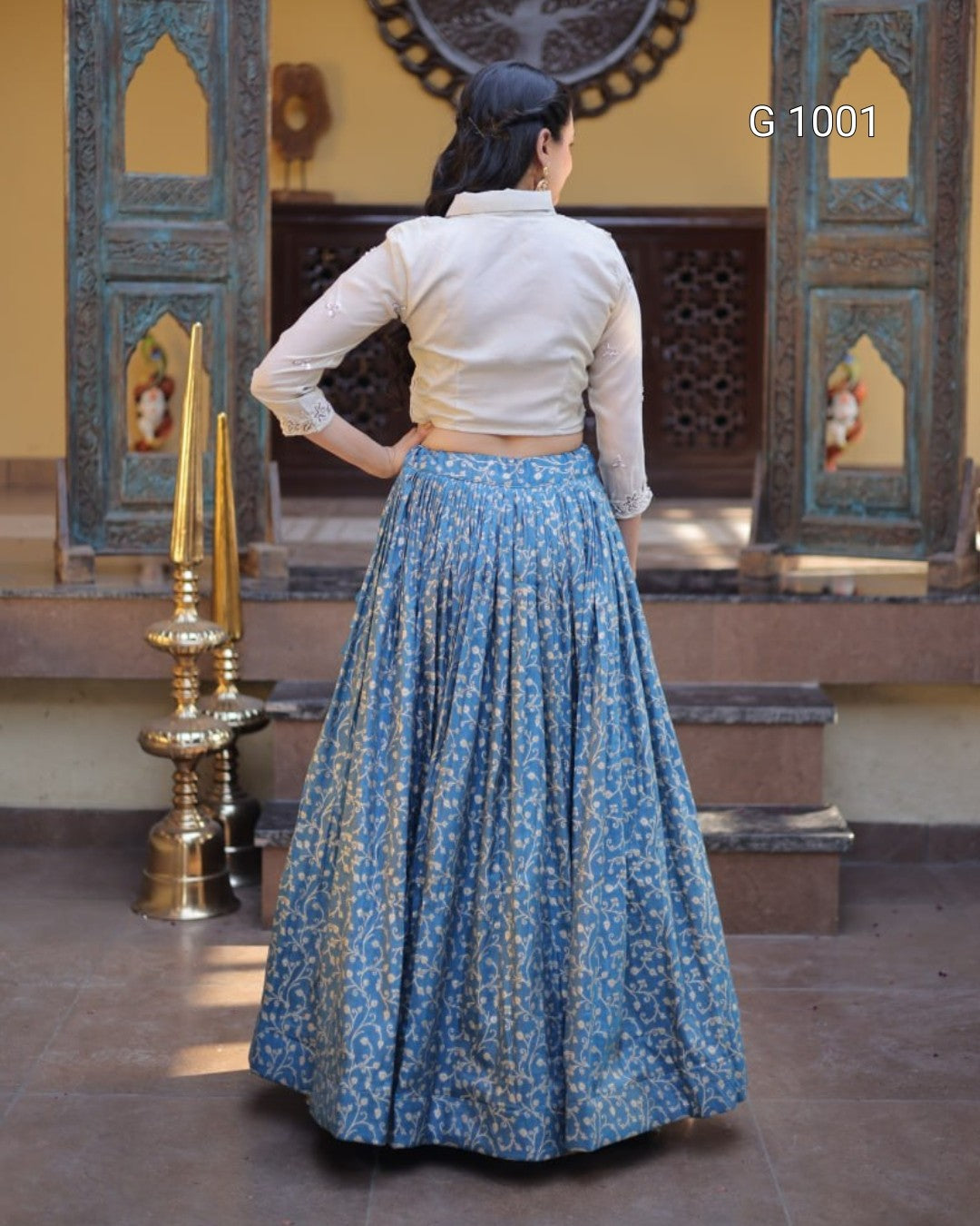 A woman stands with her back to the camera wearing a cream-colored long-sleeve crop top and a full blue patterned skirt, posed in front of an ornate doorway. The text 'G 1001' is visible in the top right corner.