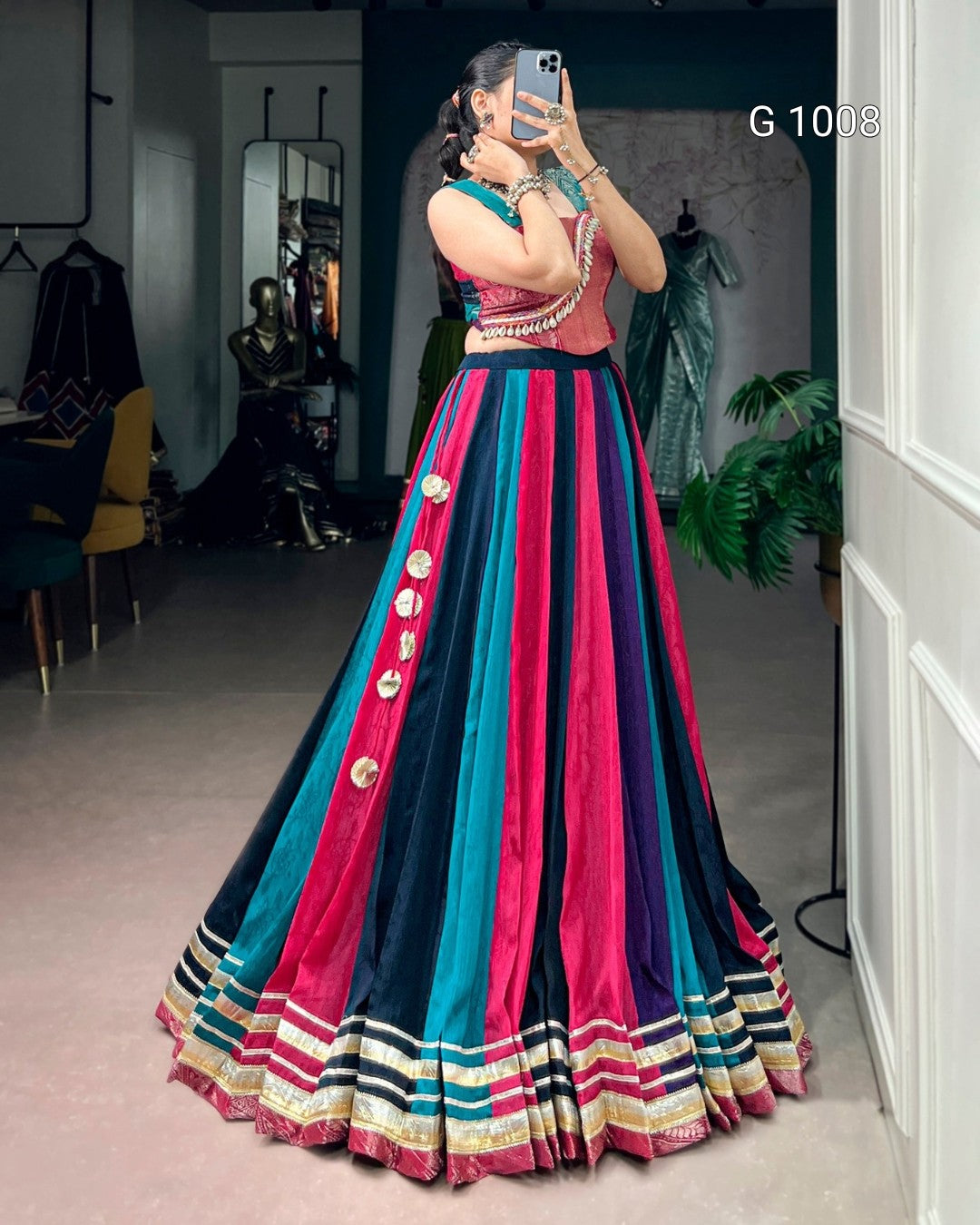 A woman takes a mirror selfie in a boutique wearing a traditional Indian lehenga choli. The skirt is made of vertical stripes of navy blue, teal, and magenta pink, adorned with gold buttons down the side and a striped border.