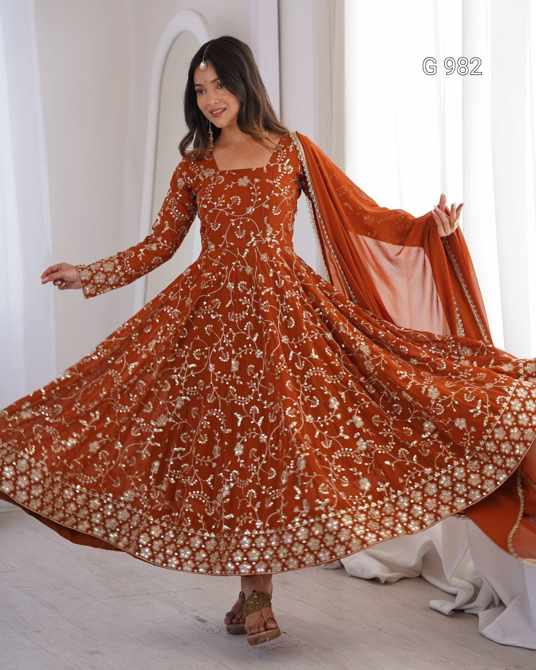 A woman modeling a rust-orange long-sleeved anarkali dress with intricate gold embroidery and a matching dupatta, captured in a bright indoor setting.