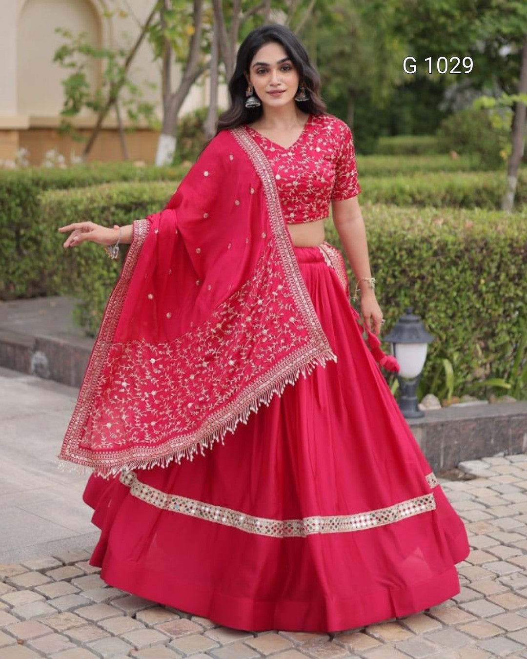 A woman modeling a bright red Indian ethnic wear set consisting of a flared lehenga skirt, a short-sleeved embroidered blouse, and a matching dupatta with gold mirror work, set against a garden background.