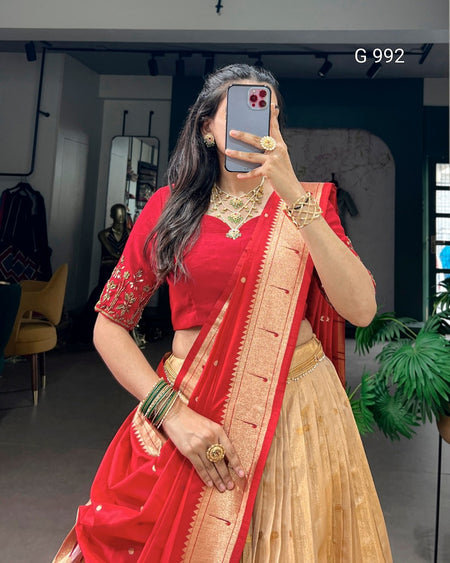 A woman takes a mirror selfie wearing a traditional Indian outfit consisting of a bright red blouse with embroidered sleeves, a matching red dupatta with a gold border, and a beige pleated skirt. She is accessorized with gold jewelry, including a necklace, rings, and bangles.