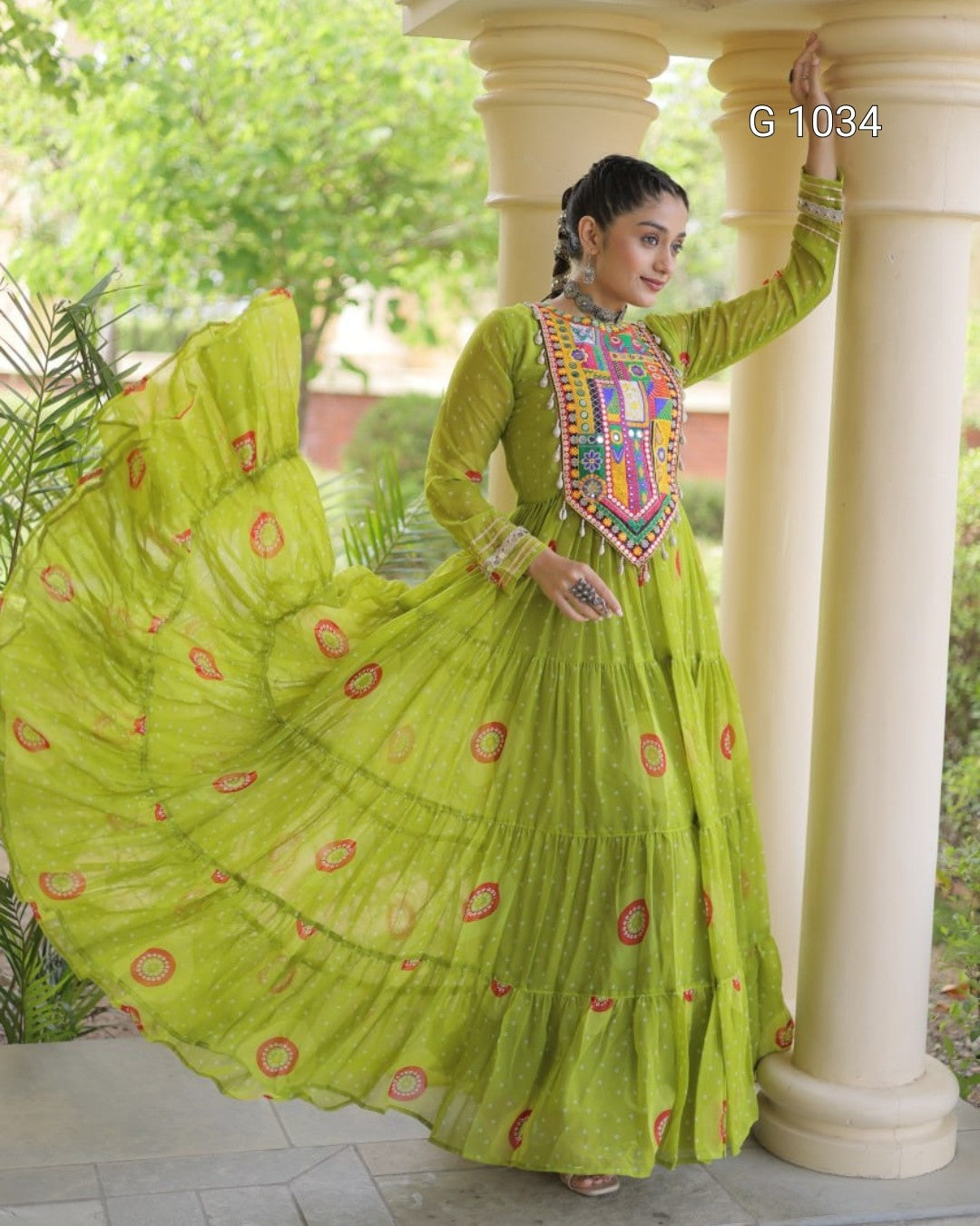 A woman modeling a bright lime green traditional Anarkali gown with a heavily embroidered multicolor yoke and a matching tiered dupatta with red circular motifs, posed in front of white pillars.