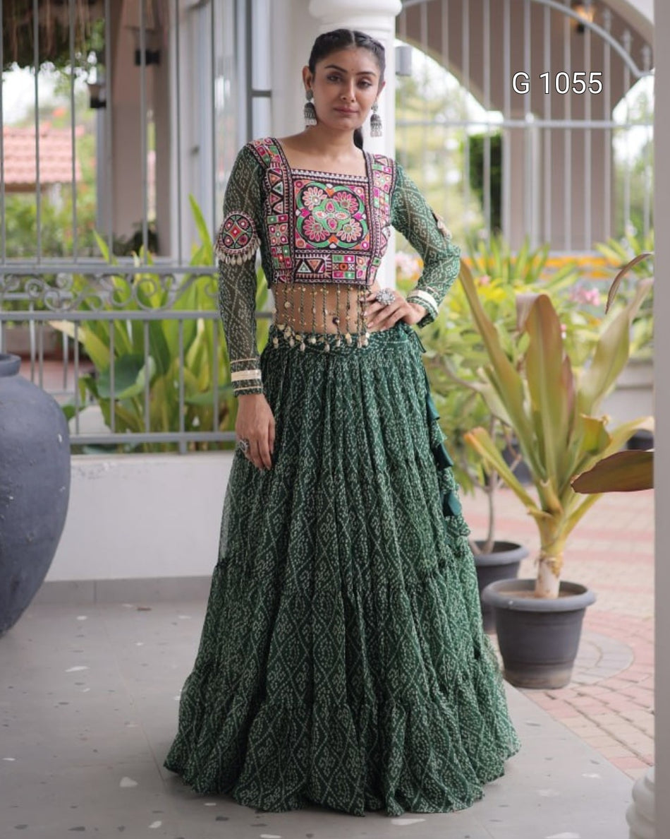 A woman models a traditional Indian ethnic wear set consisting of a dark green heavily embroidered long kurta and a matching tiered flared skirt. The kurta features a square neckline with a colorful floral mandarin embroidery in pink, red, and gold, paired with a gold-toned embellished waistband.