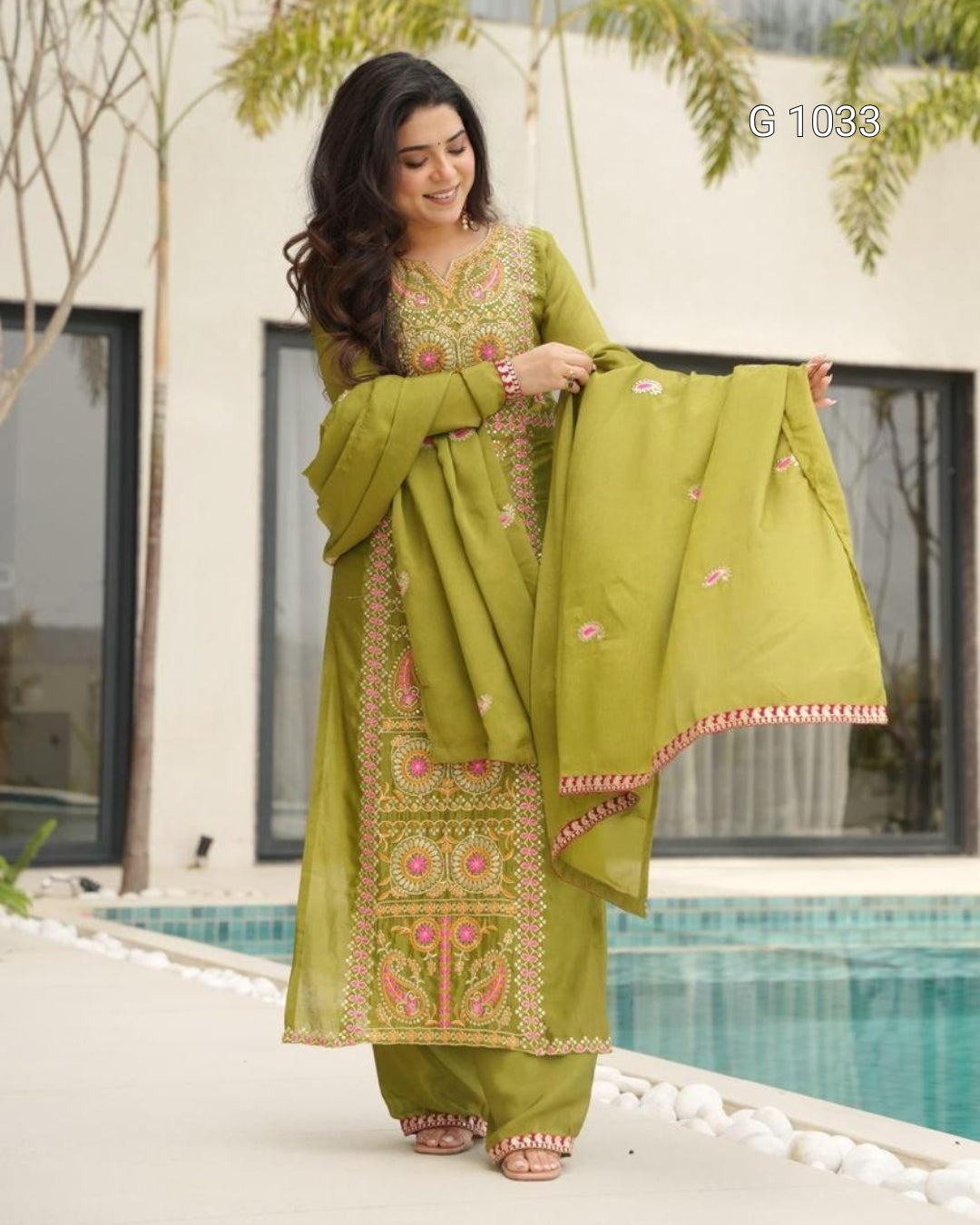 A woman models a traditional olive green ethnic salwar kameez suit with pink and gold embroidery, holding up the matching dupatta in front of a modern building and swimming pool.