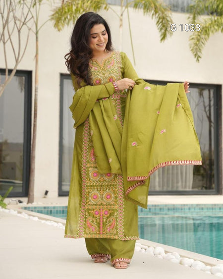 A woman models a traditional olive green ethnic salwar kameez suit with pink and gold embroidery, holding up the matching dupatta in front of a modern building and swimming pool.