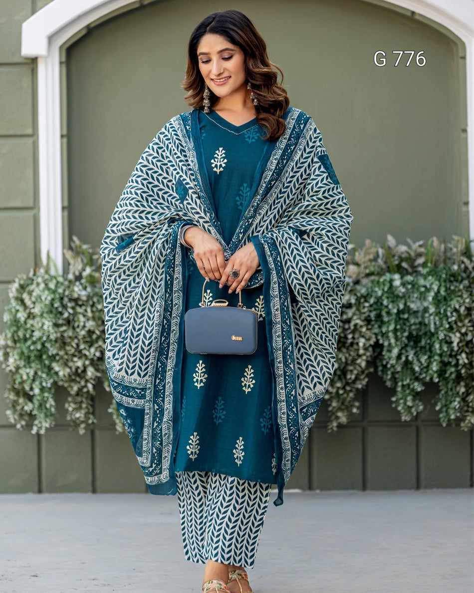 Woman wearing a teal embroidered outfit with a patterned shawl against a green wall.