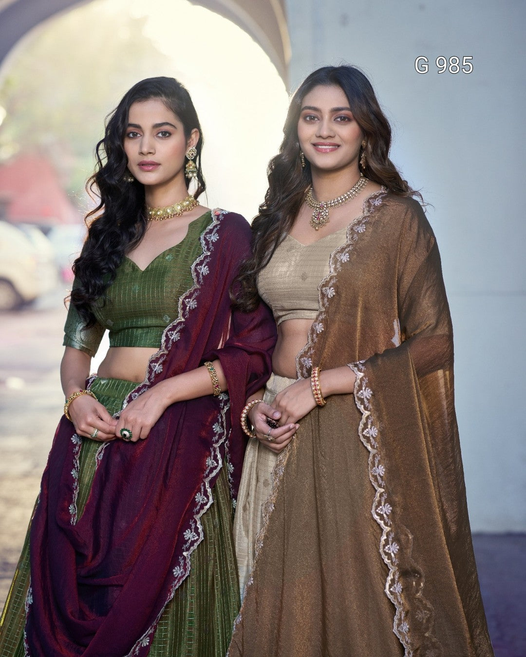 Two women modeling traditional Indian ethnic wear, standing side by side. The woman on the left wears an olive green and deep maroon lehenga choli set with silver embroidered borders, while the woman on the right wears a beige and brown set with similar silver borders.