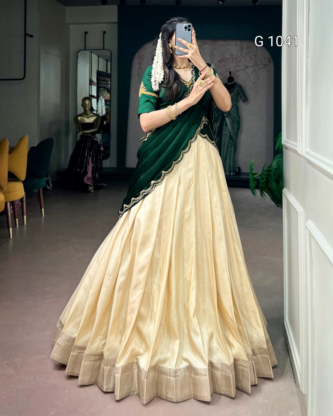 A woman takes a mirror selfie wearing a traditional Indian lehenga choli outfit. The outfit features a voluminous cream-colored skirt (lehenga) with a gold border, paired with a dark green sleeveless top (choli) and a matching dark green dupatta draped over her shoulders.