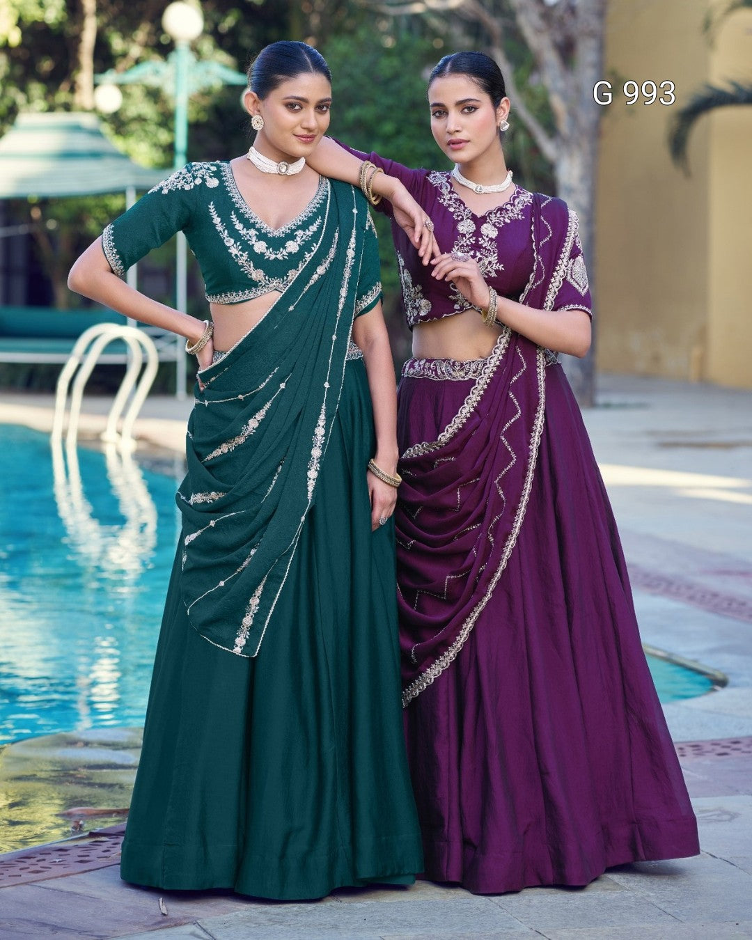 Two women modeling traditional Indian ethnic wear by a swimming pool. The woman on the left wears a dark green lehenga choli with silver embroidery, and the woman on the right wears a deep purple lehenga choli with silver embroidery.