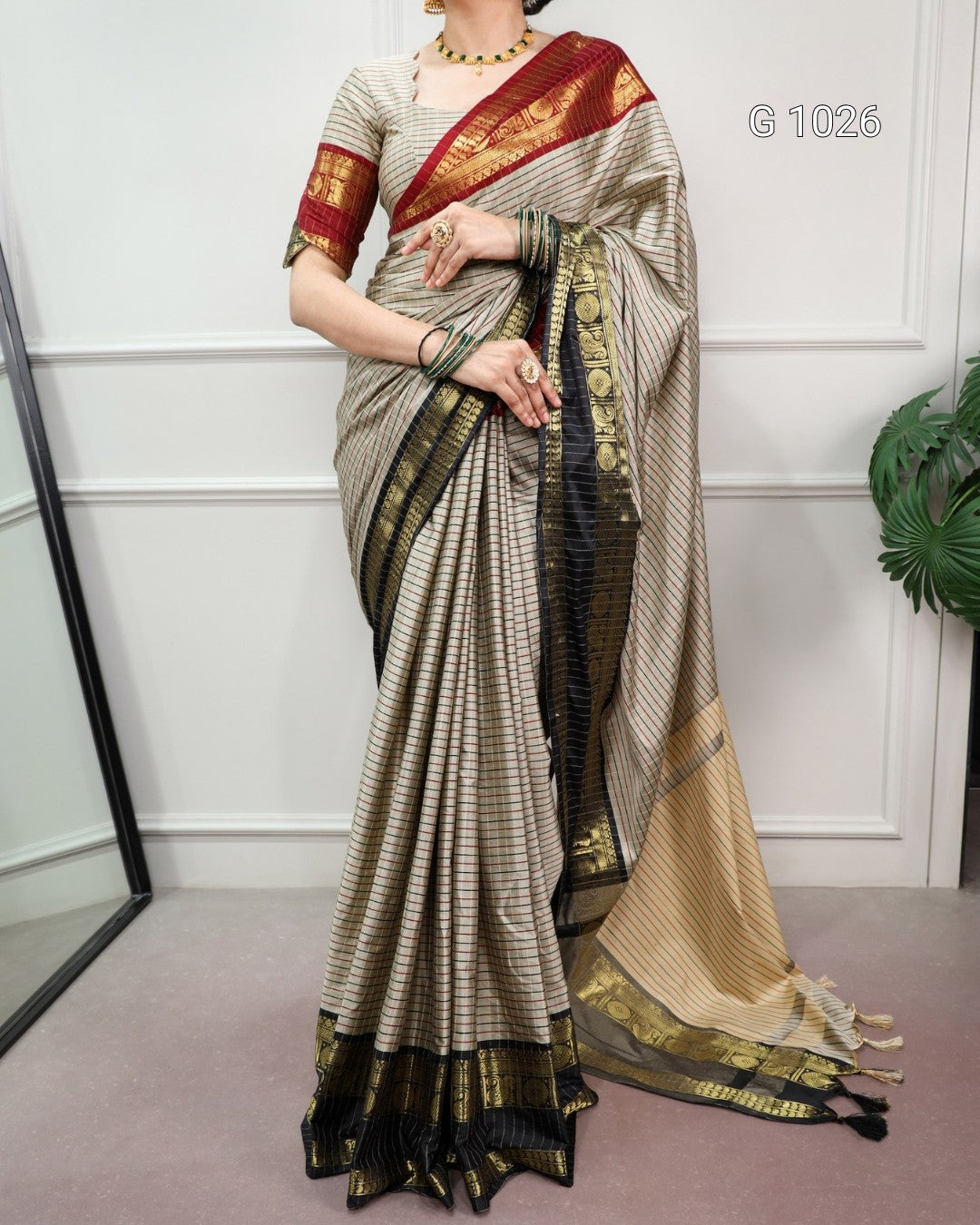 A woman models a traditional Indian saree with a beige and black striped body, a deep red border with gold motifs, and a beige pallu with a black and gold border. She is standing in an indoor setting with a white paneled wall and a plant in the background.