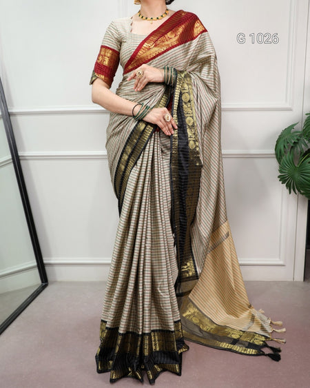 A woman models a traditional Indian saree with a beige and black striped body, a deep red border with gold motifs, and a beige pallu with a black and gold border. She is standing in an indoor setting with a white paneled wall and a plant in the background.