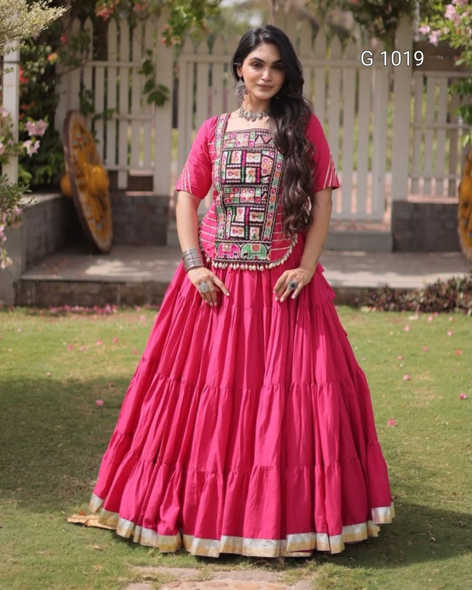 A woman wearing a bright pink traditional Indian dress with a heavily embroidered bodice and tiered skirt, standing outdoors on a grassy lawn in front of a white fence.