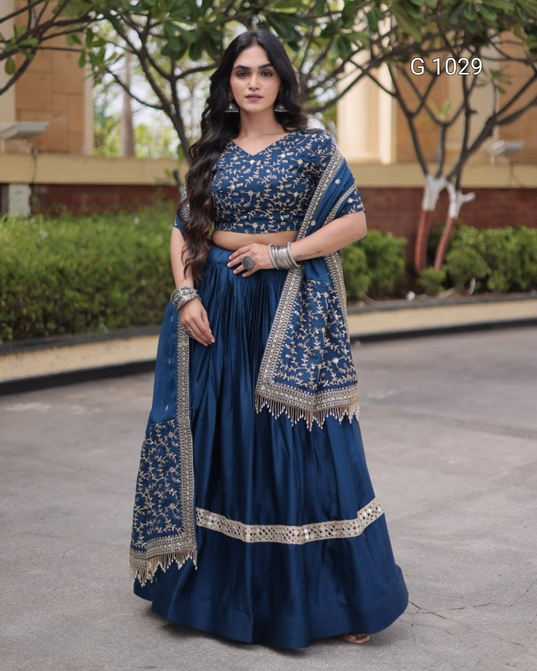 A woman modeling a navy blue traditional Indian lehenga choli set with intricate gold embroidery and a matching dupatta, standing outdoors on a paved walkway.
