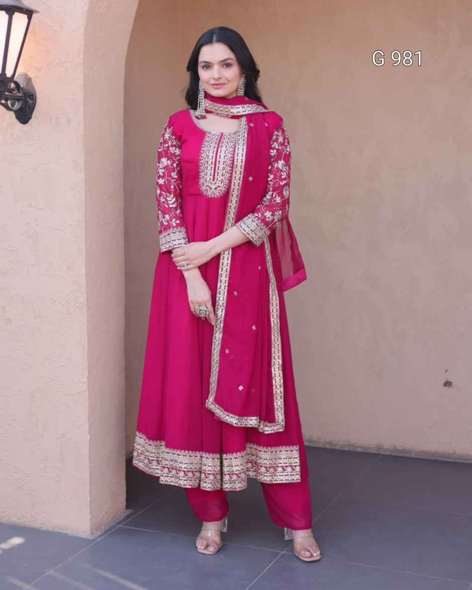 A woman modeling a bright pink traditional Indian outfit with gold embroidery, standing in front of a beige stucco wall. The outfit includes a long kurta with floral embroidery, matching pants, and a dupatta.