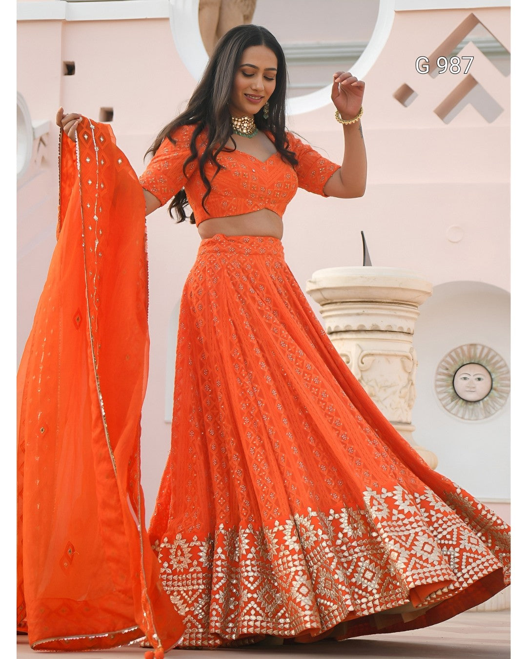An orange embroidered lehenga choli set with a matching dupatta, modeled by a woman in front of a light-colored architectural background. The outfit features intricate gold and silver embroidery, with a wide decorative border on the skirt.