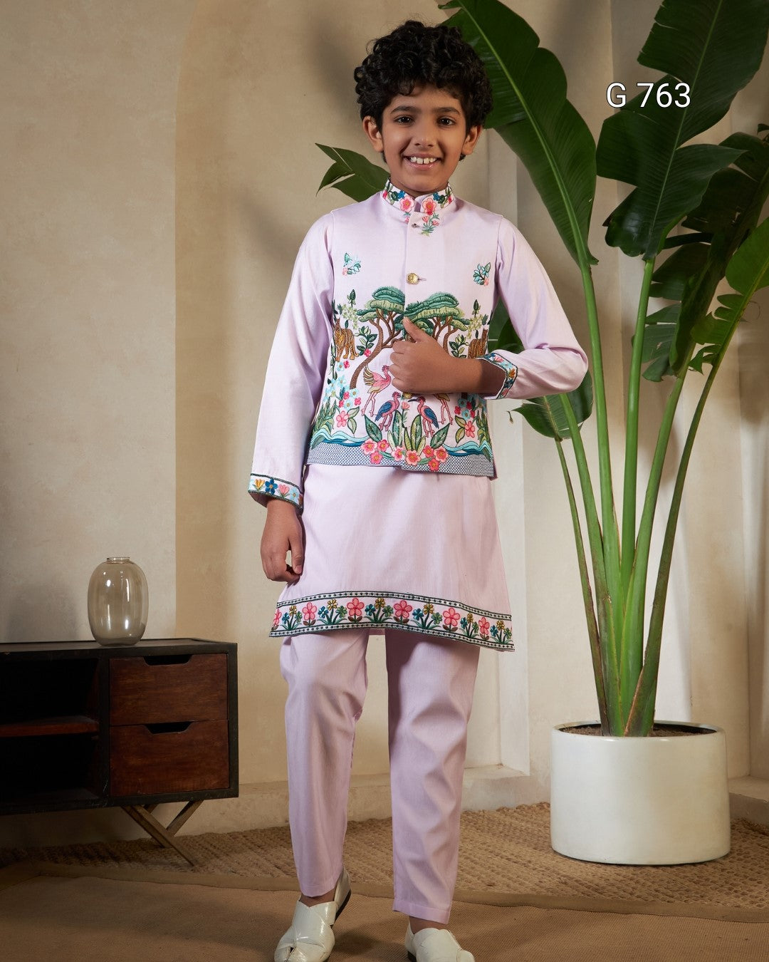 Child wearing a pink traditional outfit with floral patterns in a room with a plant and wooden cabinet.