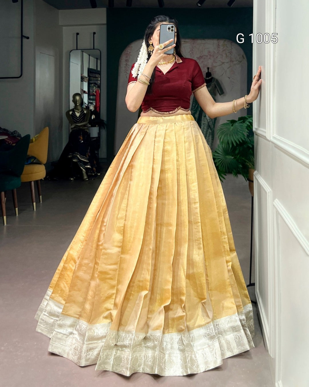 A woman takes a mirror selfie wearing a traditional Indian outfit consisting of a maroon short-sleeve blouse and a voluminous golden-yellow flared skirt with a silver border. The background shows a mirror reflecting a room with furniture.