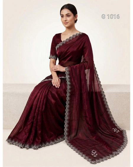 A woman seated on a beige bench wearing a deep maroon/burgundy saree with silver embroidered borders and a matching sheer dupatta. The image has a catalog number 'G1016' in the top right corner.