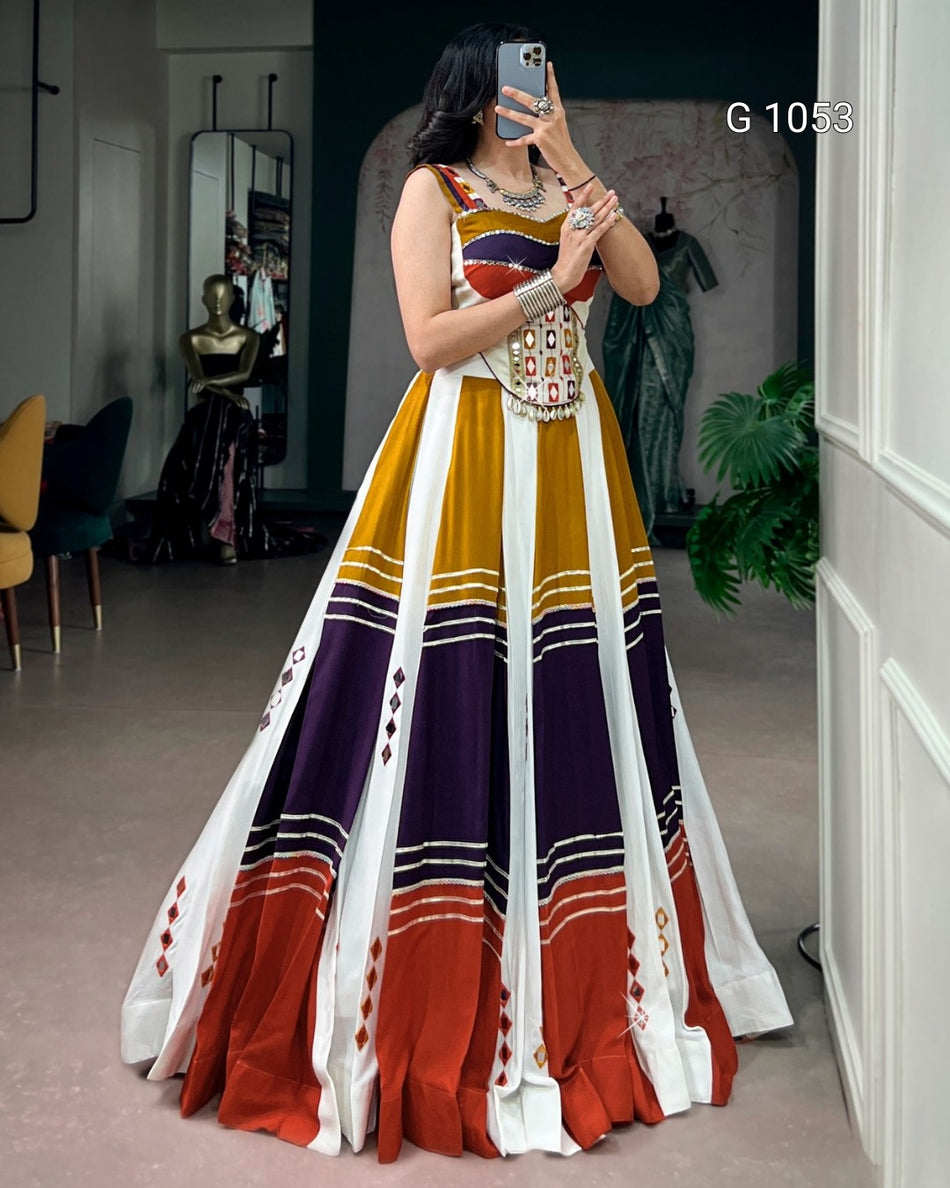 A woman takes a mirror selfie in a boutique wearing a sleeveless A-line gown with a geometric color-blocked pattern in mustard yellow, white, purple, and rust orange. The dress features a fitted bodice with a beaded waistband and a full skirt.