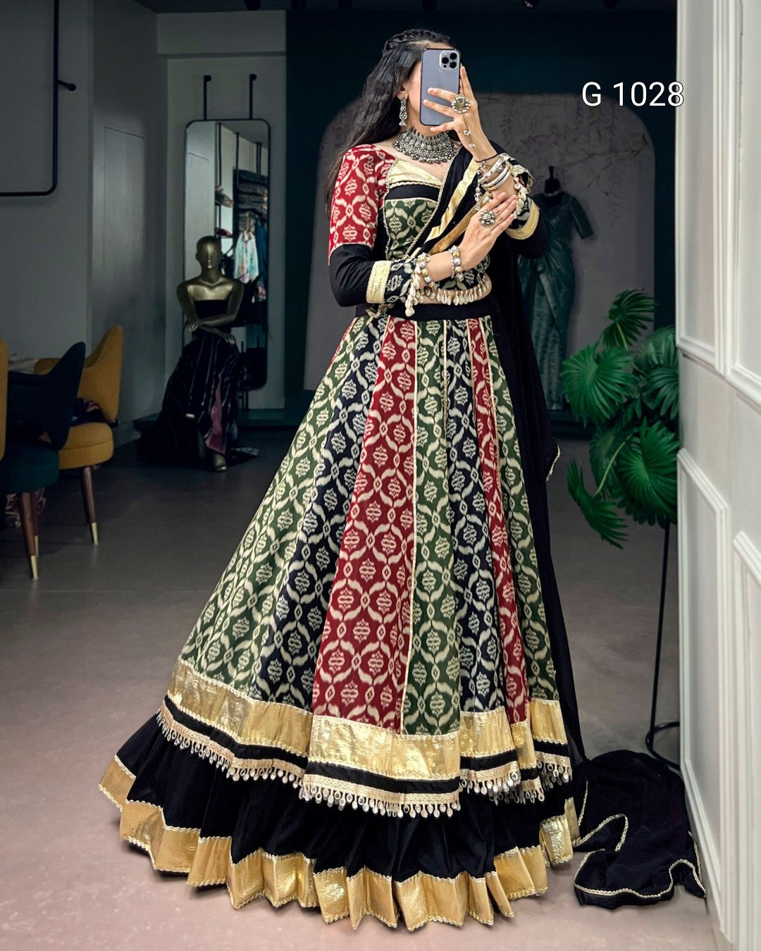 A woman takes a mirror selfie in a boutique wearing a traditional Indian lehenga choli set. The dress features vertical panels of black, red, and olive green with intricate gold and white patterns, and a black dupatta draped over her shoulder.