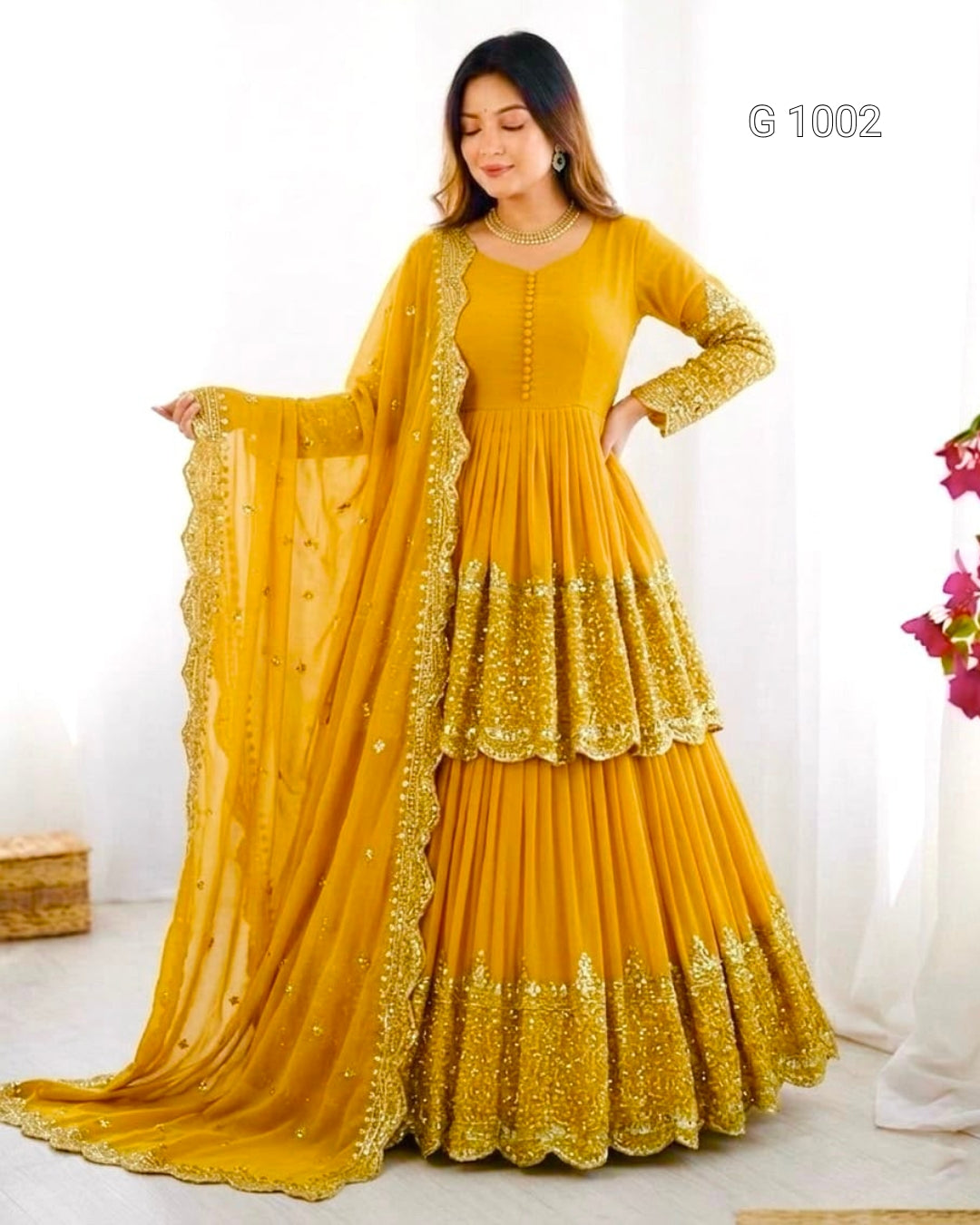 A woman modeling a traditional Indian outfit consisting of a mustard yellow long kurta with gold embroidery, a matching pleated skirt with gold border detailing, and a sheer dupatta draped over her arm. The image has a white background with decorative elements on the right side.