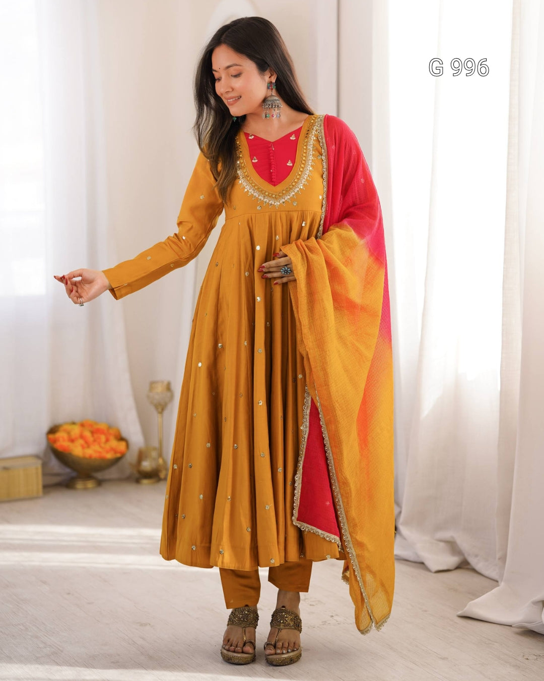 A woman models a mustard yellow Anarkali kurta set with a red inner neckline and a matching red dupatta featuring gold border detailing, set against a bright, softly lit background with white curtains.