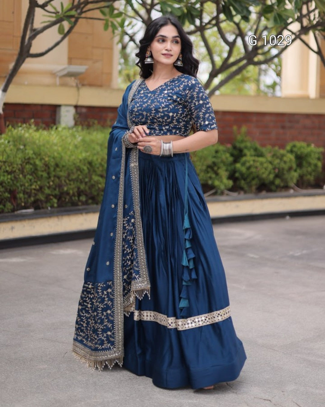 A woman models a traditional Indian navy blue lehenga choli set with intricate gold and silver floral embroidery, standing outdoors on a paved walkway with green hedges and trees in the background.