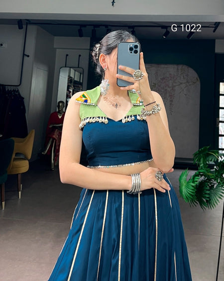 A woman takes a mirror selfie in a boutique wearing a dark teal blue two-piece outfit with a crop top featuring light green and yellow shoulder accents, a beaded neckline, and a matching pleated skirt with vertical gold stripes. She is accessorized with silver jewelry, and the background shows a clothing store interior.