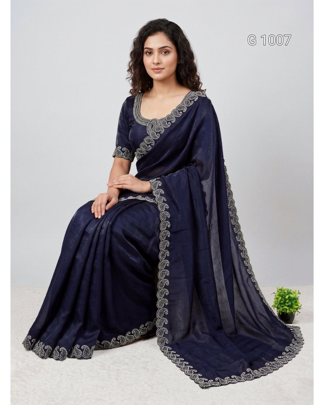 A woman seated on a light studio floor wearing a dark navy blue saree with gray embroidered borders and a matching blouse. The image has a light gray background, a small potted plant at the lower right, and the text 'G 1007' in the upper right.