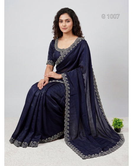 A woman seated on a light studio floor wearing a dark navy blue saree with gray embroidered borders and a matching blouse. The image has a light gray background, a small potted plant at the lower right, and the text 'G 1007' in the upper right.