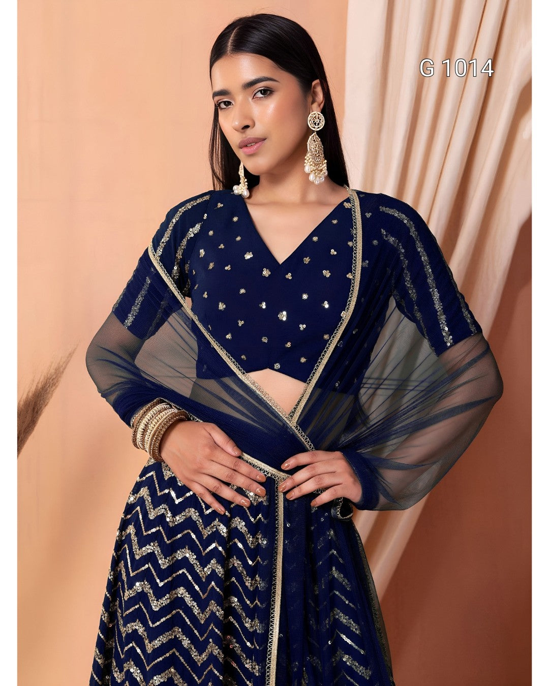 A woman models a navy blue traditional outfit with silver embellishments, featuring a patterned skirt, a decorated blouse, and a sheer matching dupatta, set against a warm beige background.