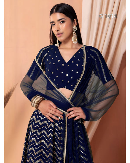 A woman models a navy blue traditional outfit with silver embellishments, featuring a patterned skirt, a decorated blouse, and a sheer matching dupatta, set against a warm beige background.