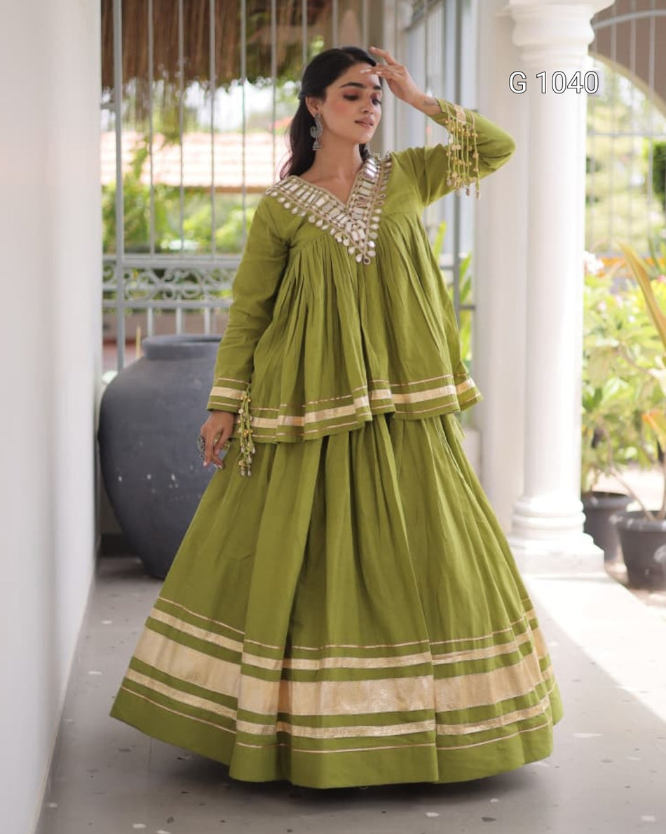 A woman models a traditional olive green Anarkali suit with gold and beige border details, standing on a covered outdoor walkway with white pillars.