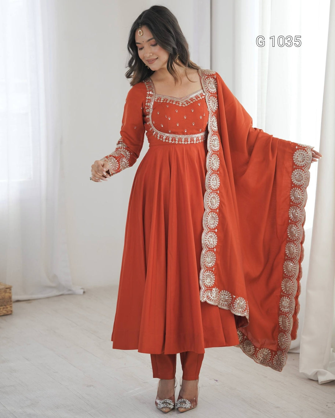A woman models a traditional Indian anarkali suit in terracotta orange with intricate silver and white embroidery, paired with matching pants and a dupatta, in a bright room with white curtains.