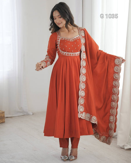 A woman models a traditional Indian anarkali suit in terracotta orange with intricate silver and white embroidery, paired with matching pants and a dupatta, in a bright room with white curtains.