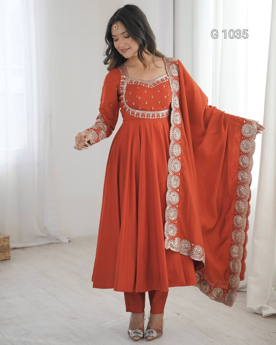 A woman models a traditional Indian anarkali suit in terracotta orange with intricate silver and white embroidery, paired with matching pants and a dupatta, in a bright room with white curtains.