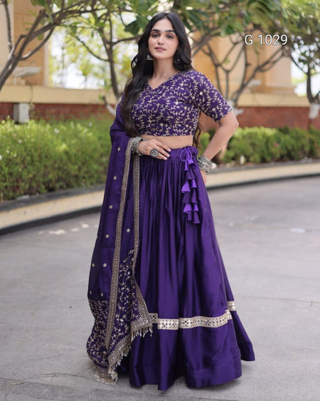 A woman modeling a purple embroidered traditional outfit with a flared skirt, embellished blouse, and draped dupatta with gold detailing, standing outdoors on a paved walkway.