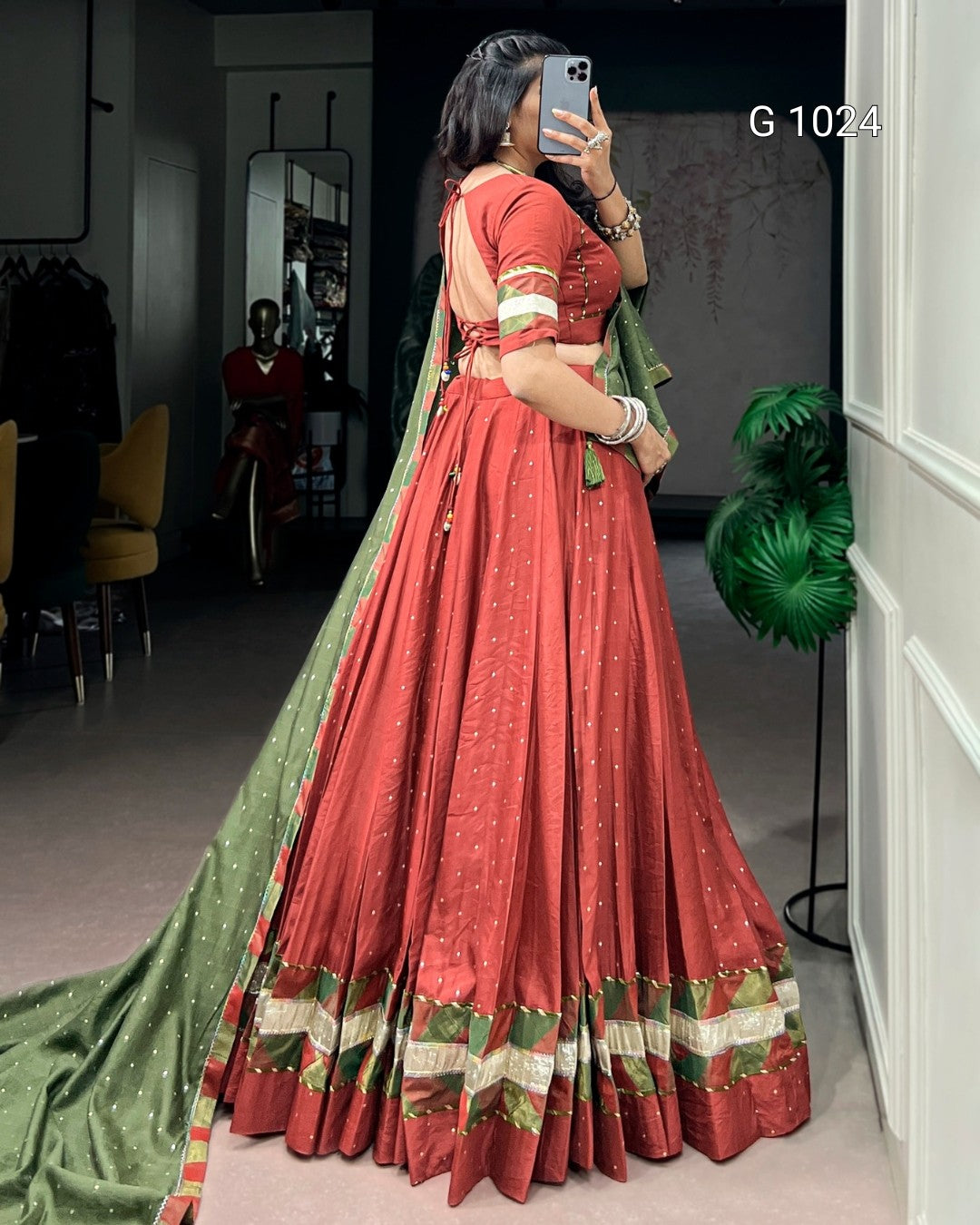 A woman takes a mirror selfie in a boutique wearing a traditional Indian red lehenga choli with a green dupatta. The lehenga has gold foil patterns and a decorative border with red, green, and gold stripes.