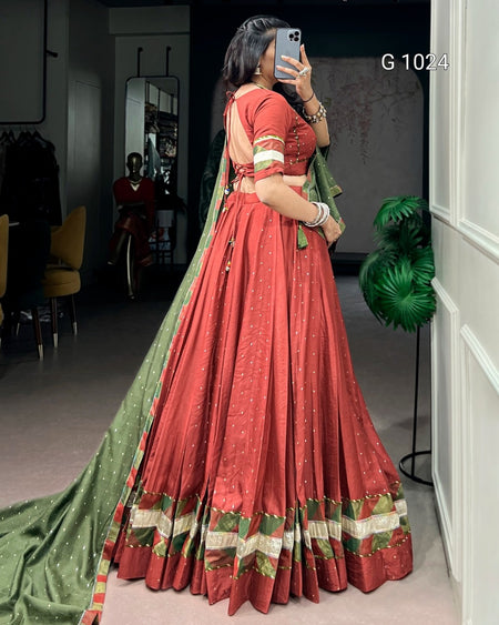 A woman takes a mirror selfie in a boutique wearing a traditional Indian red lehenga choli with a green dupatta. The lehenga has gold foil patterns and a decorative border with red, green, and gold stripes.
